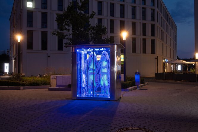 Fluoreszierende Gestalten stehen in einem Schaukasten auf einer dunklen Straße