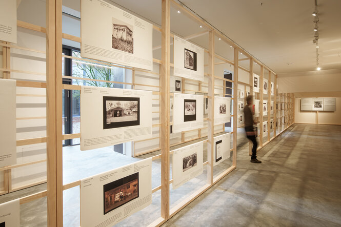 In einer Ausstellung hängen an einer hölzernen Zwischenwand historische Fotos von Gebäuden