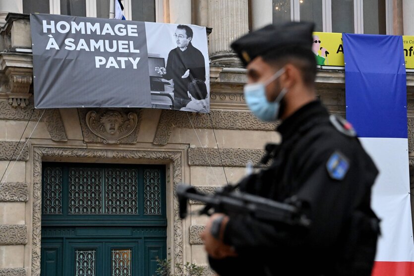 Ein Gedarm mit Maschinengewehr im Anschlag im Vordergrund. Im Hintergrund eine Gründerzeithausfassade, daran hängt ein Banner mit einem Foto von Samuel Paty und der Aufschrift "Hommage à Samuel Paty"