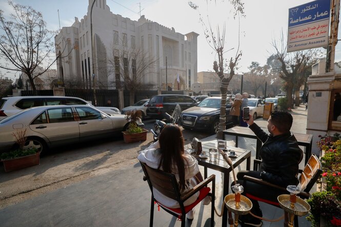 Im Vordergrund sitzt ein Pärchen an einem Tisch vor einem Restaurant. Auf der gegenüberliegenden Straßenseite das palastartige Gebäude der katarischen Botschaft. Er macht ein Handyfoto des Gebäudes.