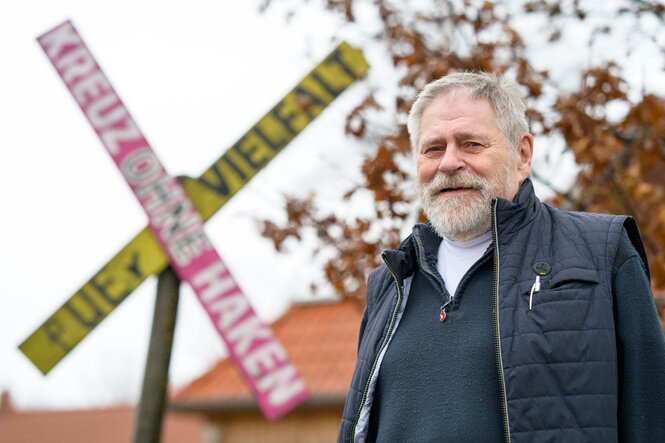 ein älterer Mann mit grauem Vollbart steht vor einem pink-gelben "X"