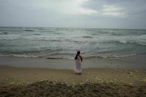 Eine Person steht am Strand und blickt auf das Meer, auf dem Boden liegen bunte Blumen verstreut