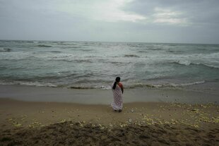 Eine Person steht am Strand und blickt auf das Meer, auf dem Boden liegen bunte Blumen verstreut