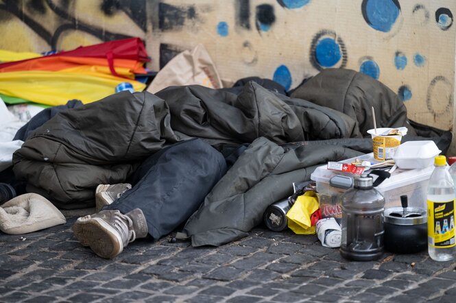 Eine offenbar wohnungslose Person liegt unter einem Schlafsack auf der Straße, daneben zahlreiche Utensilien wie Flaschen und Zigaretten