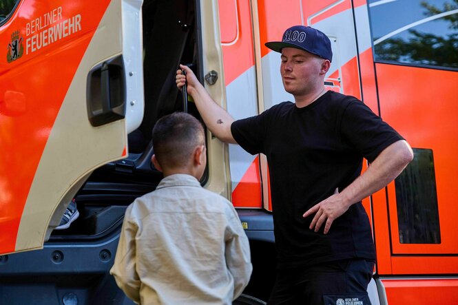 Feuerwehrmann und ein Kind vor einem Feuerwehrauto beim ersten Fußballspiel der Outreach-Jugendlichen gegen die Feuerwehr