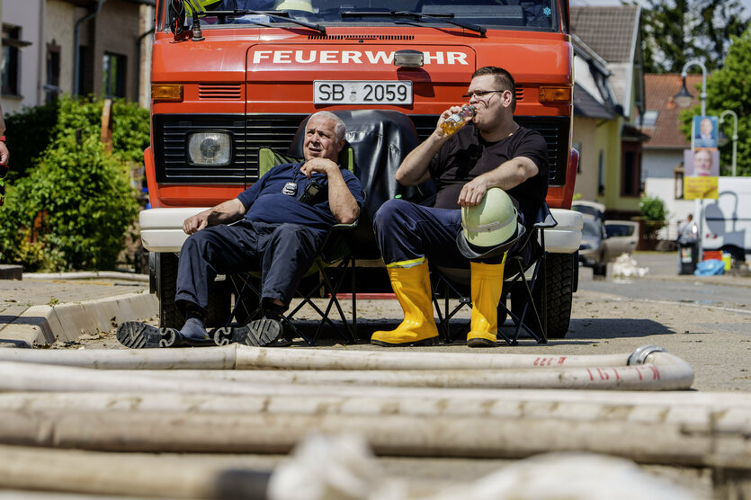 zwei Feuerwehrleute sitzen vor ihrem Fahrzeug, drumrum Flutschäden