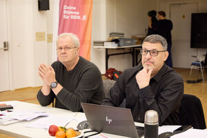 Zwei Männer sitzen an einem Tisch, dahinter ein Banner in rot-orange