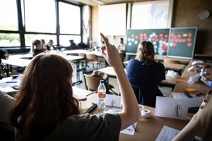 Schüler*innen sitzen im Klassenzimmer. Die Schülerin im Fordergrund meldet sich. An der Tafel hängen bunte Papiere.