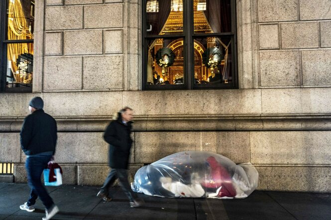 Eine Person sitzt in einem Sack aus durchsichtiger Folie auf dem Gehweg einer Straße. An ihr laufen Passanten in Winterkleidung vorbei. Oberhalb der Person sieht man von Kronleuchtern die hell erleuchteten wiesigen Fenster eines Gebäudes.