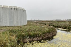 Eine graue Mauer, ein Graben, daneben eine Wiese und ein Kirchturm am Horizont