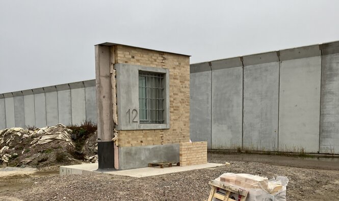 Ein Stück Wand mit Fenster vor einer hohen, grauen Mauer