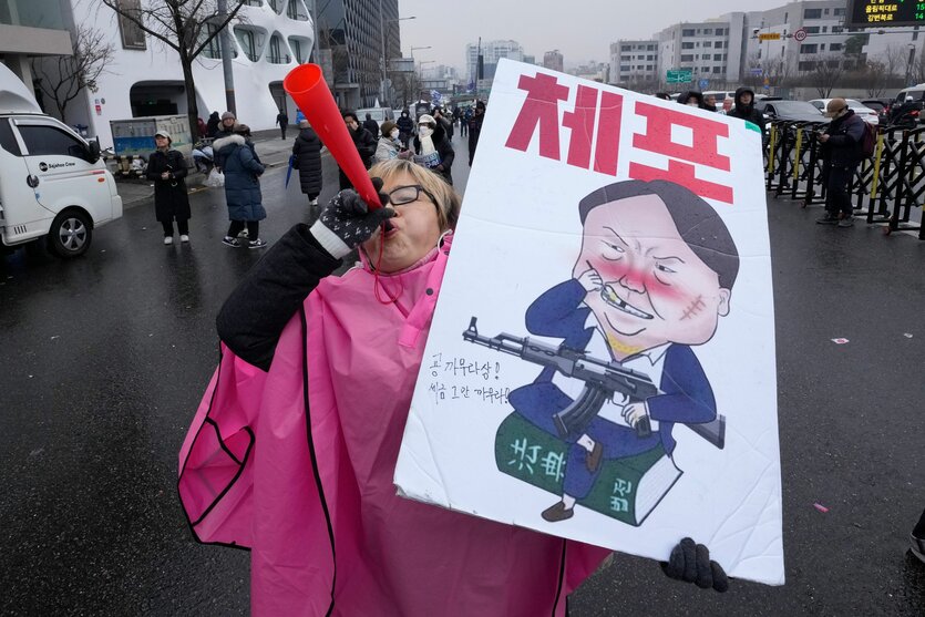 Eine Demonstrantin bläßt in einer Vuvuzela und hält ein Schild mit der Aufschrift "Verhaftet Yoon Suk Yeol". Das zeigt den suspendierten Präsidenten mit einem Sturmgewehr.