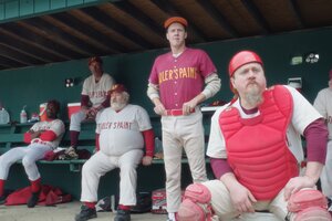 Die Mitglieder eines Baseball-Teams sitzten im Dugout-Bereich und blicken in Richtung Feld. Sie tragen weiß-rote Outfits auf denen der Schriftzug 