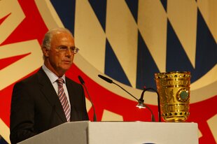 Franz Beckenbauer am Rednerpult neben dem DFB-Pokal, im Hintergrund das Vereinslogo des FC Bayern München