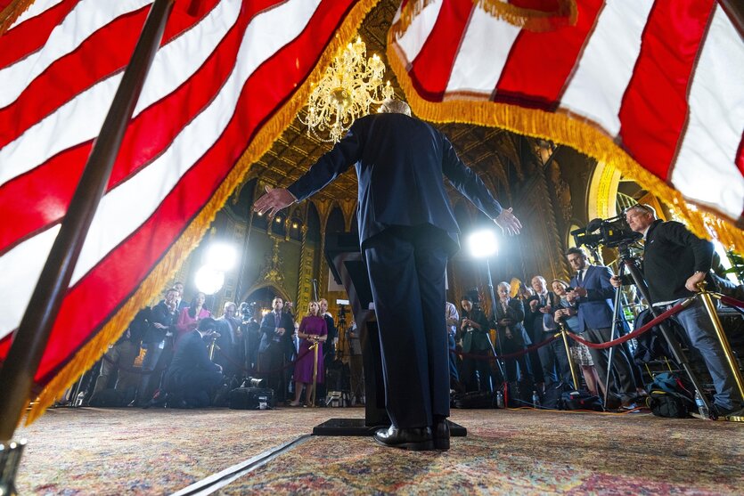 Rückansicht von Donald Trump, der vor einem Pult steht. Im Hintergrund viele Journalisten und Fotografen.