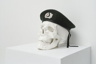 3-D-Skulptur eines Schädels auf einem Sockel. Dem Schädel ist ein Militärbarett mit Militärbrosche aufgesetzt.