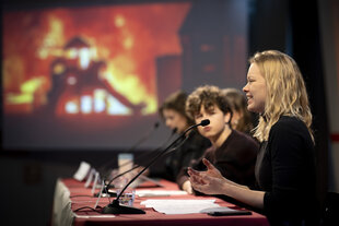 Eine junge Frau sitzt mit anderen auf einem Podium und spricht, im Hintergrund ist ein Foto eines brennenden Hauses unscharf zu erkennen