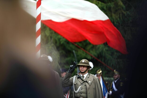 Ein Soldat mit Helm und Paradeuniform steht salutierend vor einer wehenden polnischen Flagge