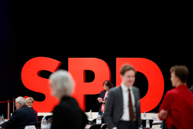 Ein großes rotes SPD-Logo in einer Halle. Davor einige Menschen.