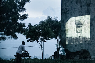 Ein Mann, nur als Uriss zu erkennen, richtet die Technik ein für eine Filmprojektion im Freien. Der Film wird an eine brückelnde Mauer projiziert