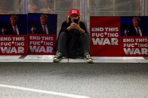 Eine Person mit roter Schirmmütze sitzt am Rande einer Straße vor Plakaten mit dem Bild Donald Trumps und der Aufschrift 