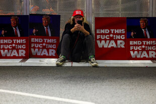 Eine Person mit roter Schirmmütze sitzt am Rande einer Straße vor Plakaten mit dem Bild Donald Trumps und der Aufschrift "End this fucking war"