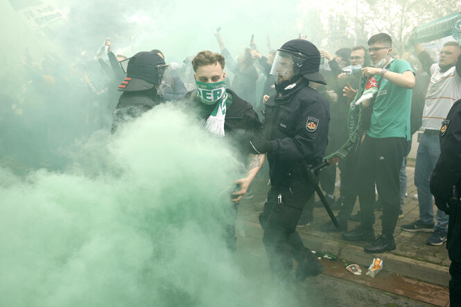 Ein mit einem Schal von Werder Bremen vermummter Stadionbesucher rangelt mit Polizisten in grünem Raum