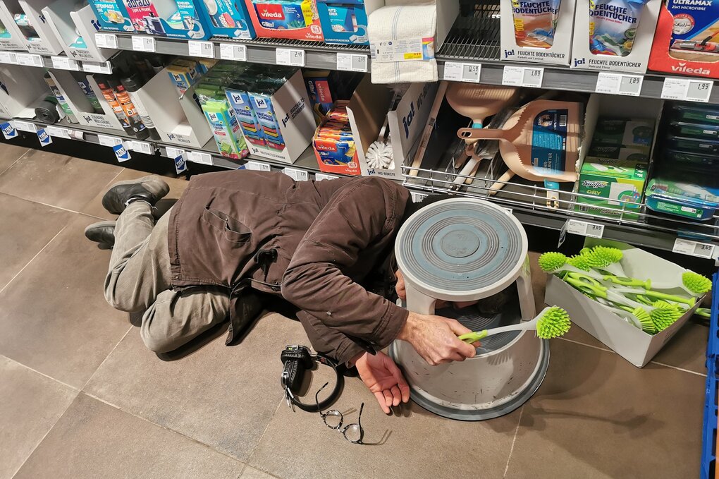 Ein Mensch liegt vor einem Supermarkt-Regal mit Reinigungsutensilien auf dem Boden. In einer Hand hält er eine Reinigungsbürste.