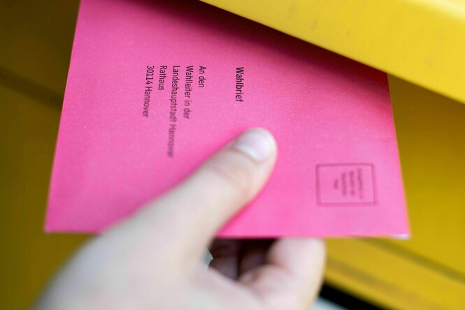 Eine Hand hält einen rosafarbenen Wahlbrief, der zur Hälfte in dem Schlitz eines gelben Briefkastens steckt