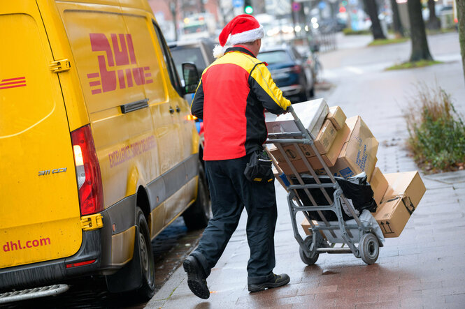 Ein Paketbote mit Weihnachtsmannmütze fährt einen Stapel Pakete auf einer Sackkarre aus