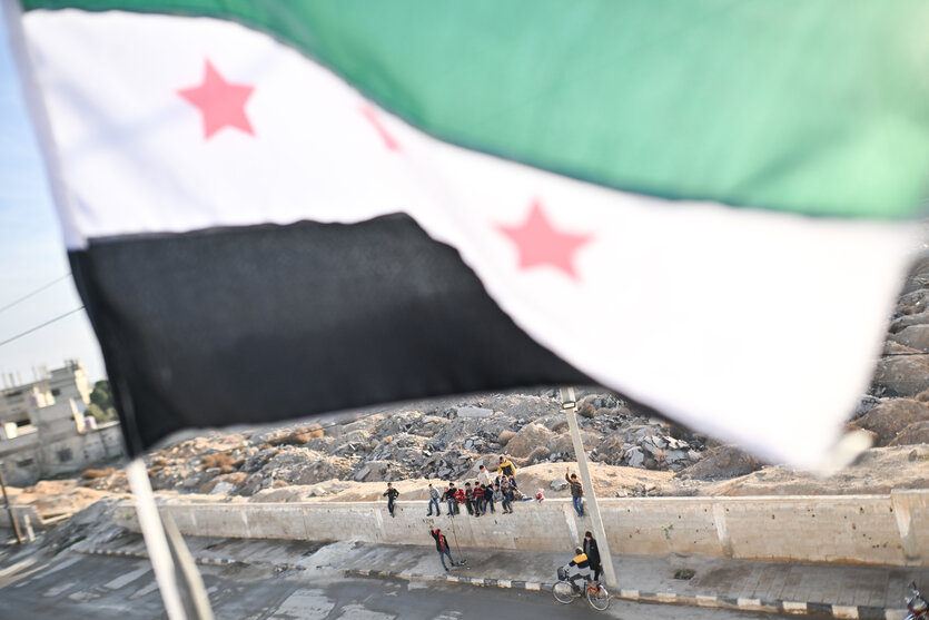 Kinder und Jugendliche sitzen auf einer Mauer. Über ihnen weht die Fahne der syrischen Revolution.