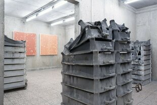 In einem Ausstellungsraum sind hohe Stapel aus Transportkisten aus Plastik aufgestellt. Die Stapel in Hell- und Dunkelgrau sind ineinander zusammengeschmolzen.