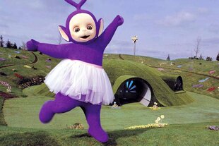 Ein Teletubby steht, bekleidet in einem Tutu, auf einem Bein vor seinem mit Gras bewachsenen Häuschen und lächelt.