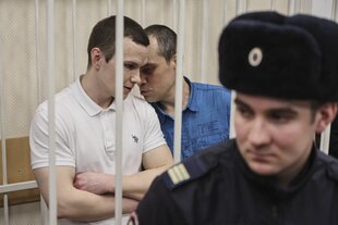 Zwei Männer tuscheln, sie sind hinter Gittern, vor ihnen steht ein russischer Polizeibeamter