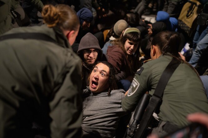 Mehrere Menschen sitzen auf dem Boden, einer von ihnen hat weit aufgerissene Augen als ihn Polizisten, die nur von hinten zu sehen sind, anpacken, um ihn fortzutragen,