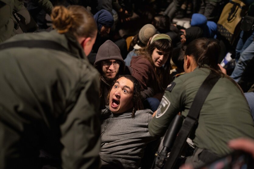 Mehrere Menschen sitzen auf dem Boden, einer von ihnen hat weit aufgerissene Augen als ihn Polizisten, die nur von hinten zu sehen sind, anpacken, um ihn fortzutragen,