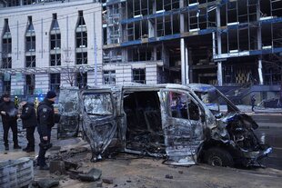 Ein zerstörtes Auto wird von Polizisten inspiziert, im Hintergrund ein beschädigtes Gebäude