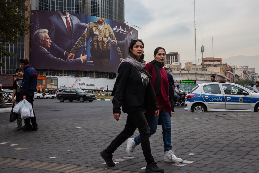 Straßenszene aus Teheran, zwei Frau gehen ohne Hijab über einer Platz, im Hintergrund Propagandaplakat gegen Israel