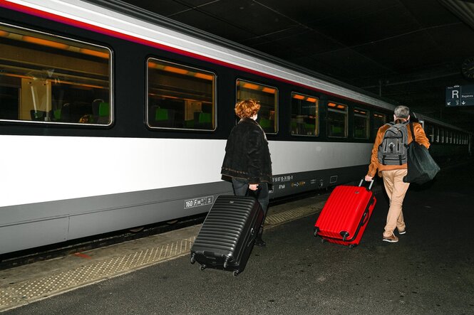 Ein Mann mit einem roten Koffer und eine Frau mit einem schwarzen Koffer gehen an einem Bahnsteig entlang