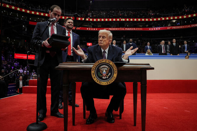 Trump sitzt in einer großen Halle gestikulierend hinter einem Schreibtisch