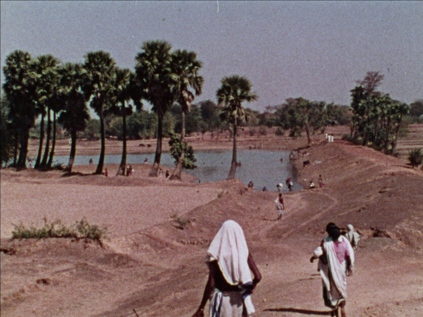 Landschaftsaufnahme in Westbengalen, die im hinteren Bildteil einen See zeigt, der von Bäumen gesäumt ist. Im vorderen Bildteile laufen Menschen in Richtung des Sees.