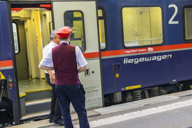 Ein Schaffner unterhält sich mit einem Kollegen, sie stehen auf dem Bahnsteig, hinter ihnen der Zug