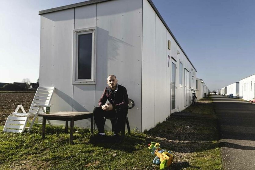 Weiße Wohncontainer Reihen auf einem Acker in Deutschland, ein Syrer sitzt wartenden von der Stirnseite und blickt uns scheinbar erschöpft an.