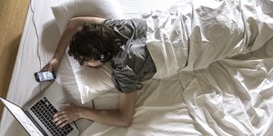 Ein junger Mann liegt mit Laptop und Smartphone im Bett