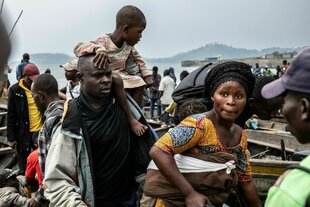 Vor einer Hügellandschaft am Seeufer an welchem geschäftigt belebte Boote angelegt, schaut eine Frau besorgt in die Kamera, hinter ihr ein Mann mit kind auf der Schulter.