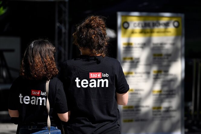 Das Bild zeigt zwei weibliche Personen von hinten. Sie tragen schwarze T-Shirts mit der Aufschrift "taz lab team". Im Hintergrund ist verschwommen ein Schild zu sehen. Der Text des Schildes ist nicht lesbar.