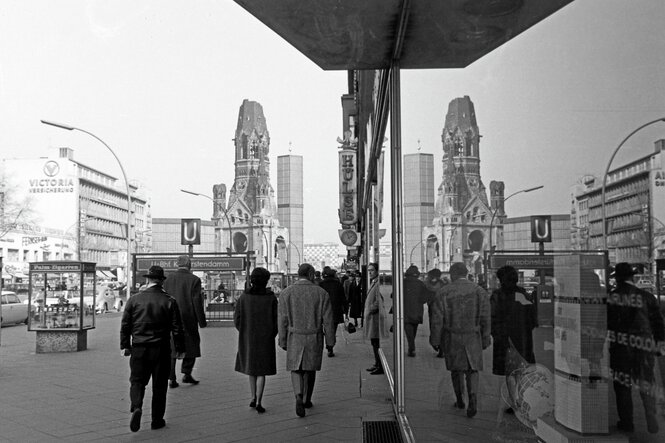 Passanten auf dem Kurfürstendamm in Berlin spiegeln sich mit der Kaiser Wilhelm Gedächtniskirche in Schaufensters