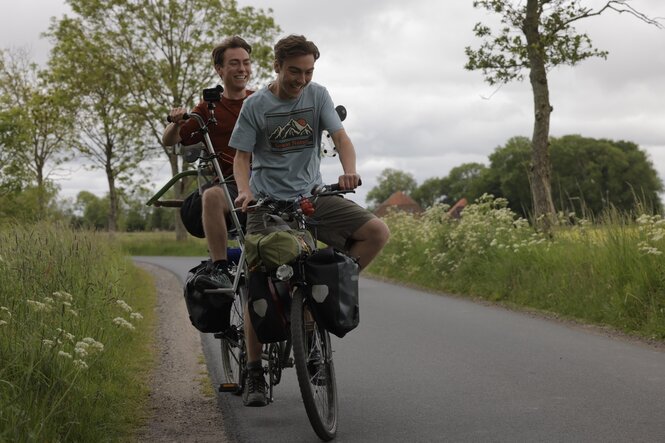 Zwei junge Männer fahren gemeinsam auf einem Fahrrad über eine Kreisstraße in norddeutsch ländlicher Umgebung.