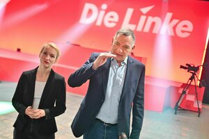 Jan van Aken und Ines Schwerdtner sprechen vor Beginn des Bundesparteitags dessen Bühne im Hintergrund zu sehen ist, Van Aken macht eine Handbewegung als würde er einen Füllstand zeigen.