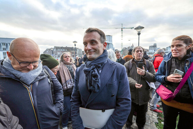 Eine Personengruppe steht vor einer urbanen Landschaft. Eine männliche Person blick betroffen zu Boden, ein Mann im Zentrum, der Ankläger, blickt ruhig nach vorne.